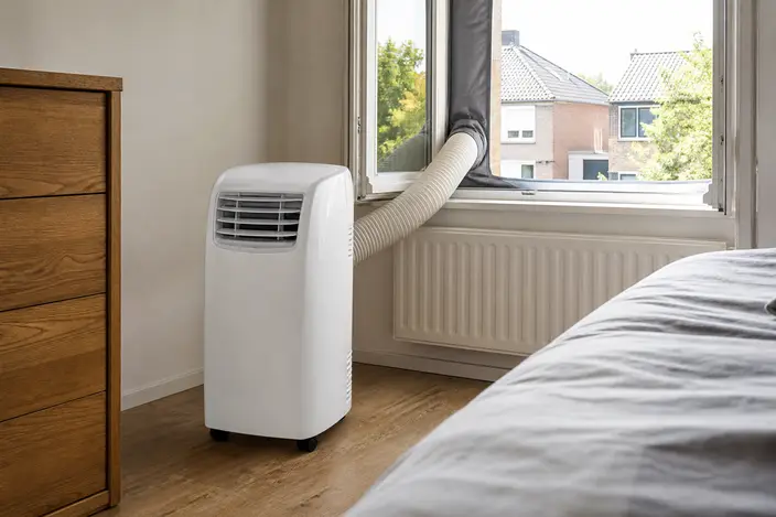 AI-beeld: een witte mobiele airco op wieltjes in een slaapkamer, met een slang naar een open raam met een raamafdichting